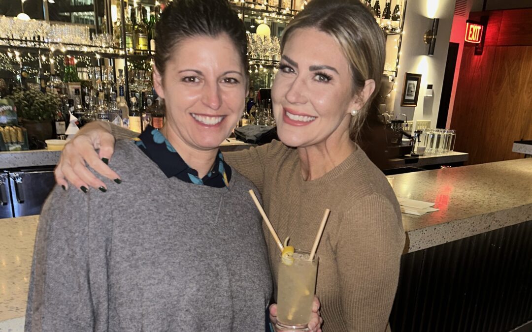 Two women stand in front of a bar smiling and holding a drink
